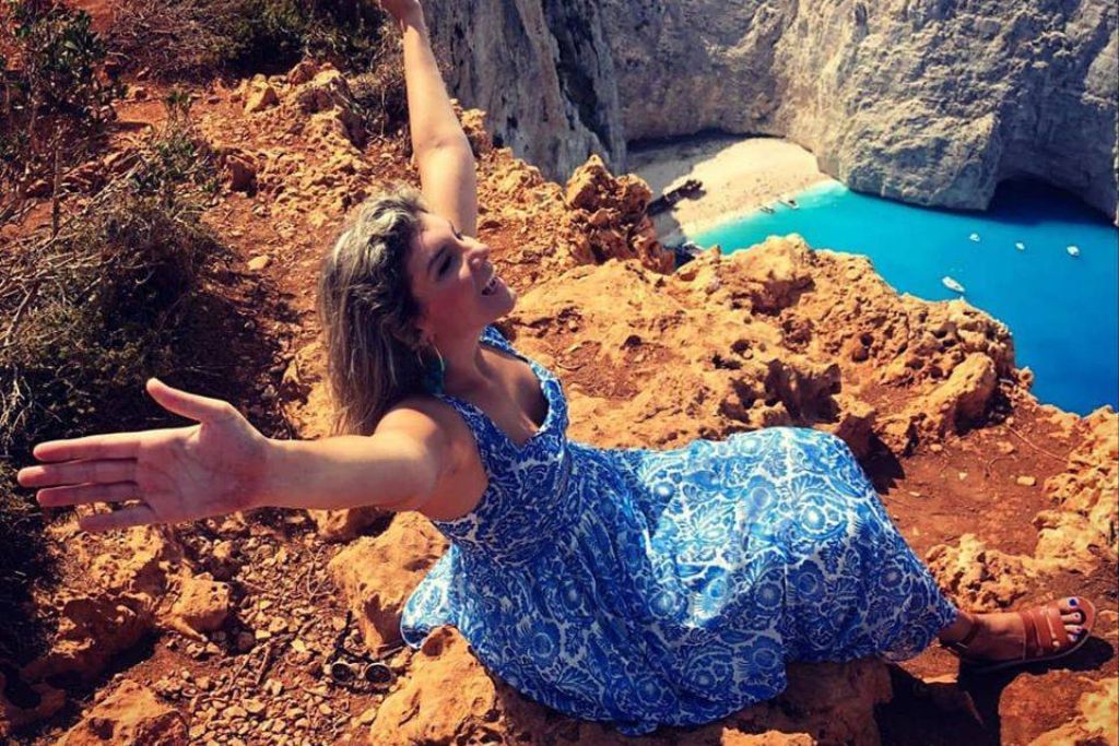 Γυναίκα με μπλε φόρεμα και ανοιχτά χέρια φωτογραφίζεται πάνω στα βράχια και από κάτω φαίνεται η θάλασσα