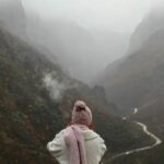 Μια κυρία ντυμένη με χειμωνιάτικα ρούχα κοιτάει προς τα βουνά και στο βάθος υπάρχει ομίχλη