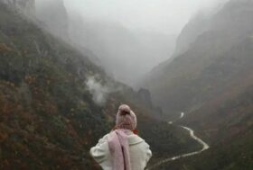 Μια κυρία ντυμένη με χειμωνιάτικα ρούχα κοιτάει προς τα βουνά και στο βάθος υπάρχει ομίχλη