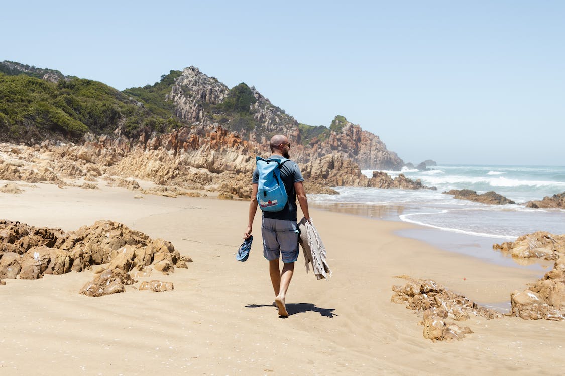 Άνδρας με σακίδιο περπατά στην άμμο μιας παραλίας και στο βάθος φαίνονται βράχια