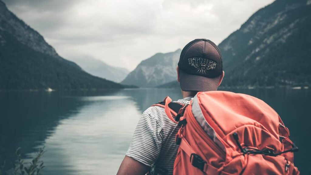 Άνδρας που φοράει κόκκινο σακίδιο και μαύρο καπέλο και είναι σε ένα ποτάμι ή λίμνη και βλέπει τη θέα και τα βουνά τριγύρω