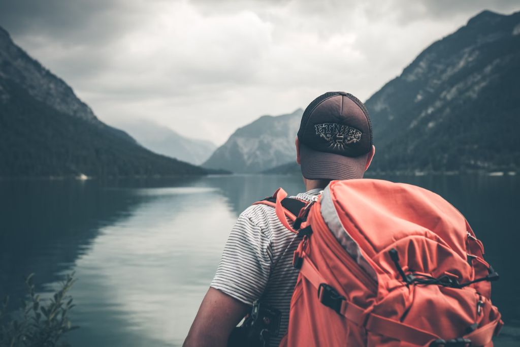 Άνδρας που φοράει κόκκινο σακίδιο και μαύρο καπέλο και είναι σε ένα ποτάμι ή λίμνη και βλέπει τη θέα και τα βουνά τριγύρω