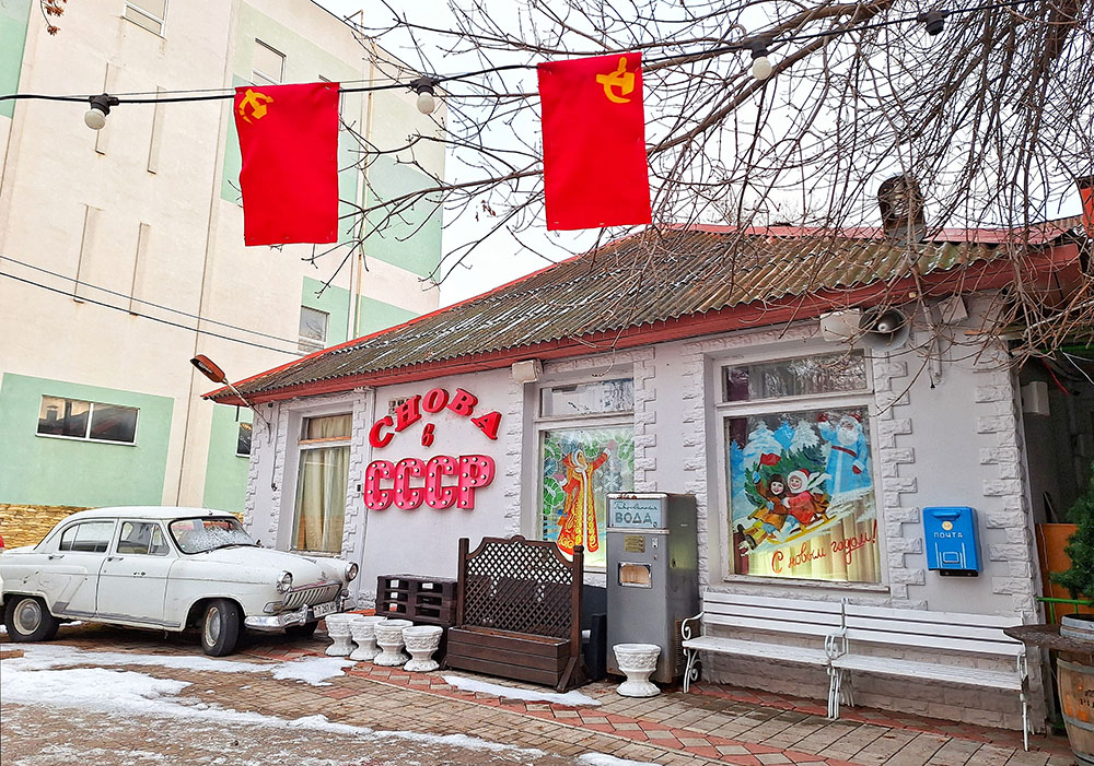 Ένα λευκό σπίτι με τρεις τζαμαρίες, λευκό παλιό αυτοκίνητο απ'έξω και κρεμασμένες κόκκινες σημαίες της Σοβιετικής Ένωσης