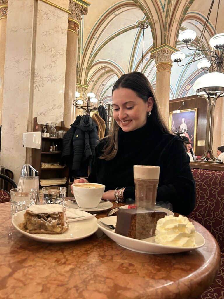 Κοπέλα που κάθεται σε ένα κοσμικό καφέ φορώντας μαύρο ζιβάγκο και στο τραπέζι έχει γλυκά και δύο καφέδες και στην άλλη φωτογραφία πιάτο με ένα κομμάτι σοκολατίνα και σαντιγύ
