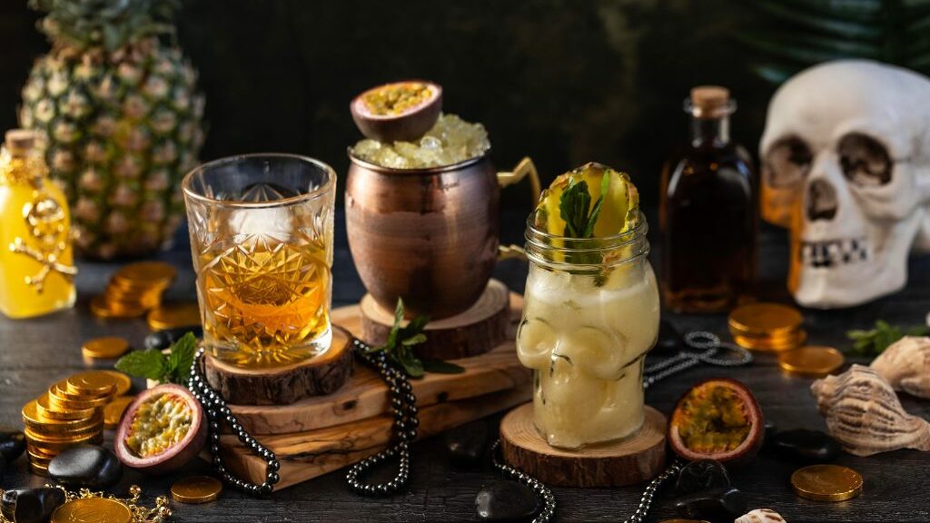 Διάφορα ποτήρια με cocktails, μια νεκροκεφαλή και ένας ανανάς πάνω σε ένα μαύρο τραπέζι