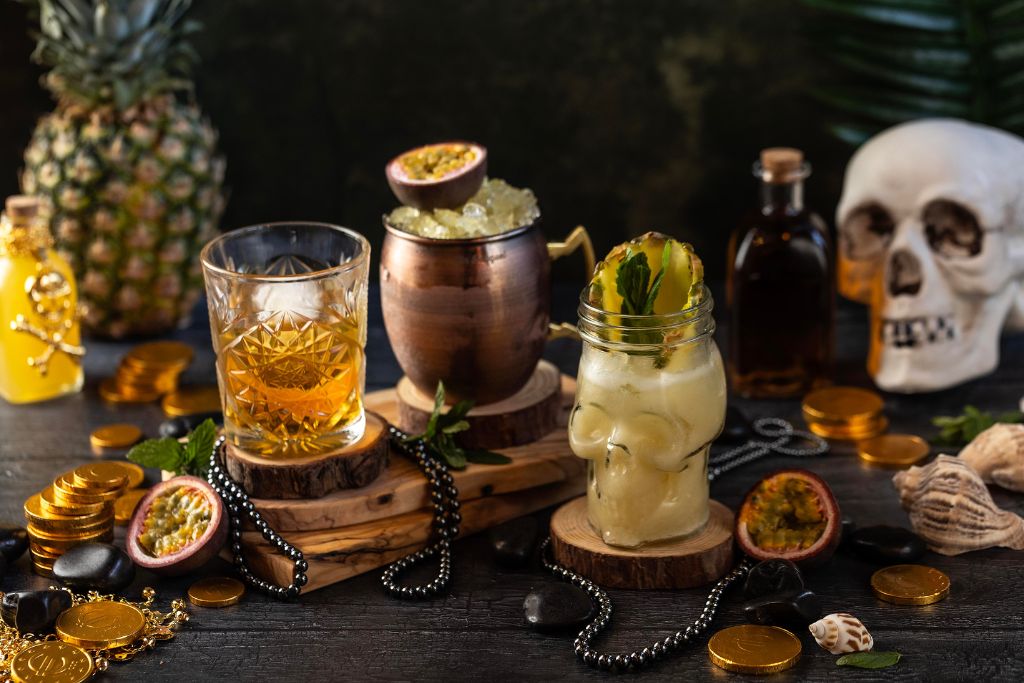 Διάφορα ποτήρια με cocktails, μια νεκροκεφαλή και ένας ανανάς πάνω σε ένα μαύρο τραπέζι