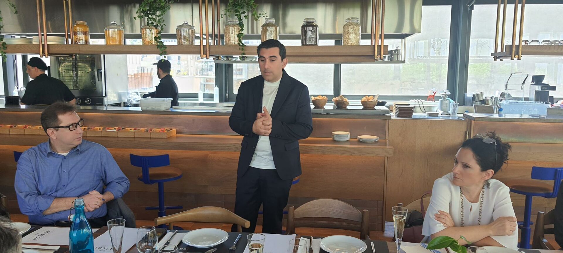 Kύριος με κοστουμι μιλάει σε κόσμο μπροστά από ένα τραπέζι με πιάτα