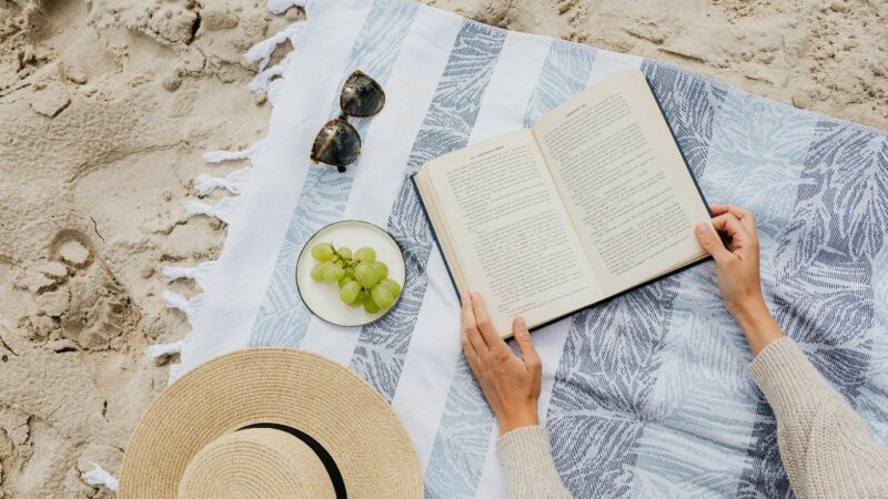 Βιβλίο πάνω σε πετσέτα θαλάσσης, άμμος από κάτω και γυαλιά ηλίου