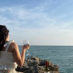 Κοπέλα που φαίνεται η πλάτη της να κρατάει ένα ποτήρι κόκκινο κρασί μπροστά στη θάλασσα