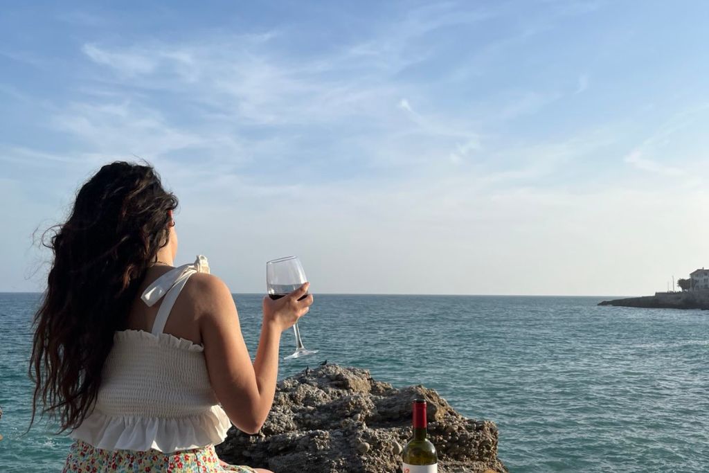 Κοπέλα που φαίνεται η πλάτη της να κρατάει ένα ποτήρι κόκκινο κρασί μπροστά στη θάλασσα