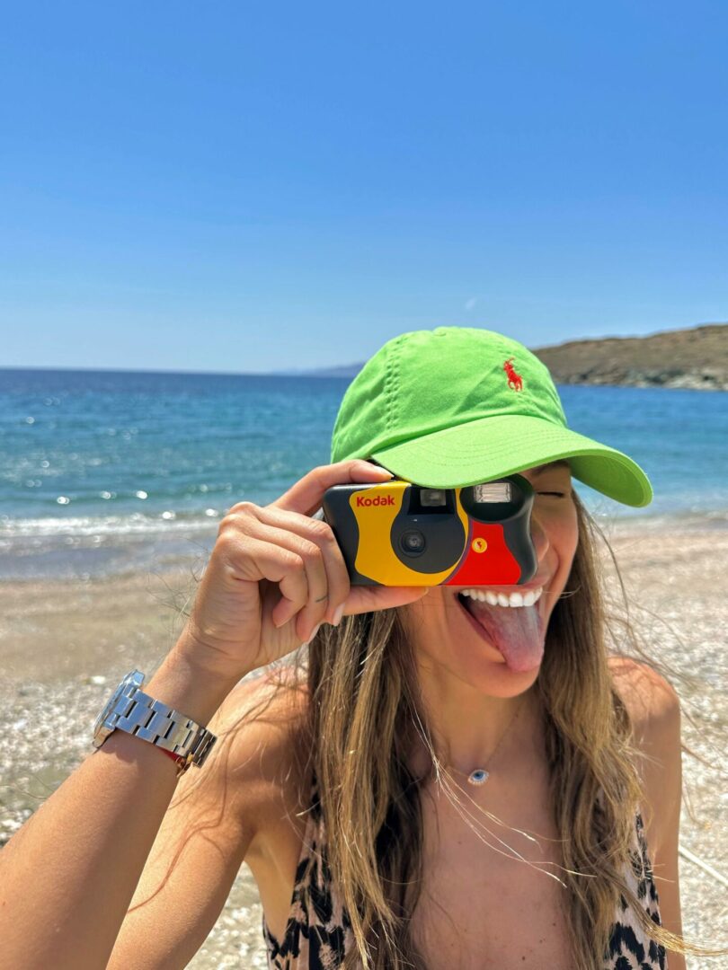 Κοπέλα με πράσινο καπέλο και φωτογραφική μηχανή μπροστά στο ένα μάτι που βγάζει τη γλώσσα σαν να ποζάρει και πίσω η θάλασσα