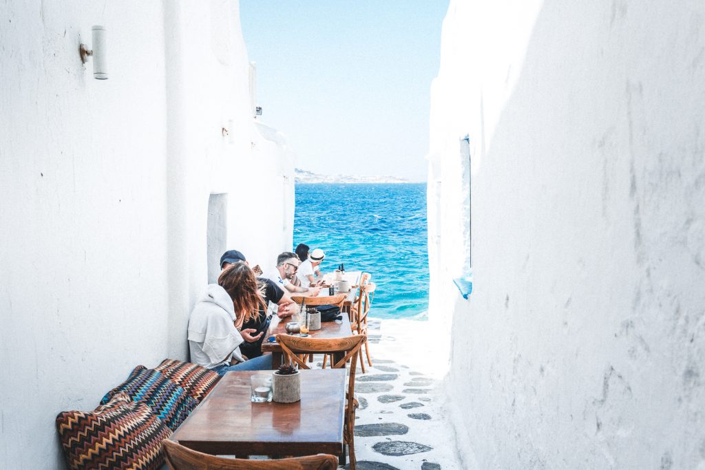 Στενό δρομάκι με λευκά σπιτάκια, τραπέζια με κόσμο και θέα στη θάλασσα