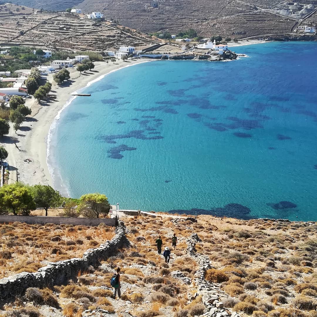 Παραλία με αρμυρίκια, λευκά σπιτάκια και κόσμος που κατεβαίνει προς αυτήν από τον λόφο